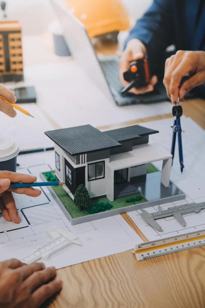 Architects' hands around a 3D house model on blueprints, using pencils and drafting tools.